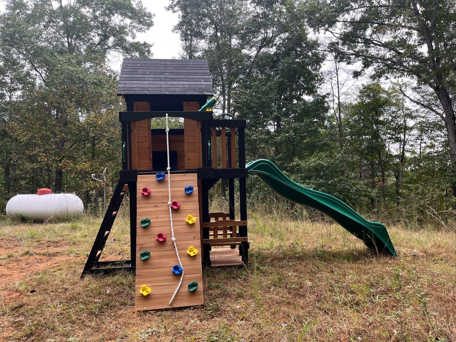 Playset climbing wall with colored rock holds and a rope beside a green wave slide in Blue Ridge, GA